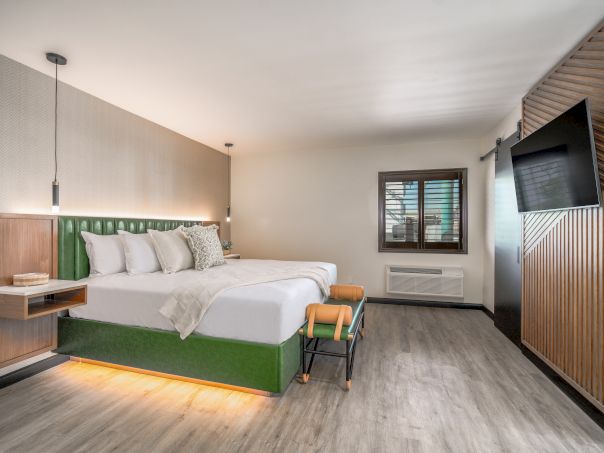 Modern hotel room with a green-bed frame, white bedding, floating headboard shelves, a wall-mounted TV, wooden accents, and a window.