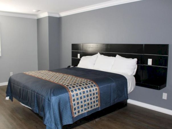A modern hotel-style bedroom with a large dark wood headboard, king bed, white pillows, blue bedding, and a gray-blue wall background. The room has a window with blinds and hardwood flooring.