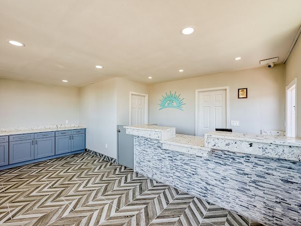 A bright reception area with a stone-tile front desk, chevron-patterned floor, light gray walls, blue-gray cabinets along left, and ocean-themed wall decor.