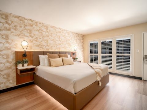 A modern bedroom with a large bed, decorative wall, bedside lamps, plants, and windows with shutters fills the space.