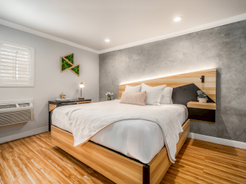 A modern bedroom with a wooden platform bed, gray accent wall, white bedding, bedside shelves, a small desk by the window, and warm wood flooring.