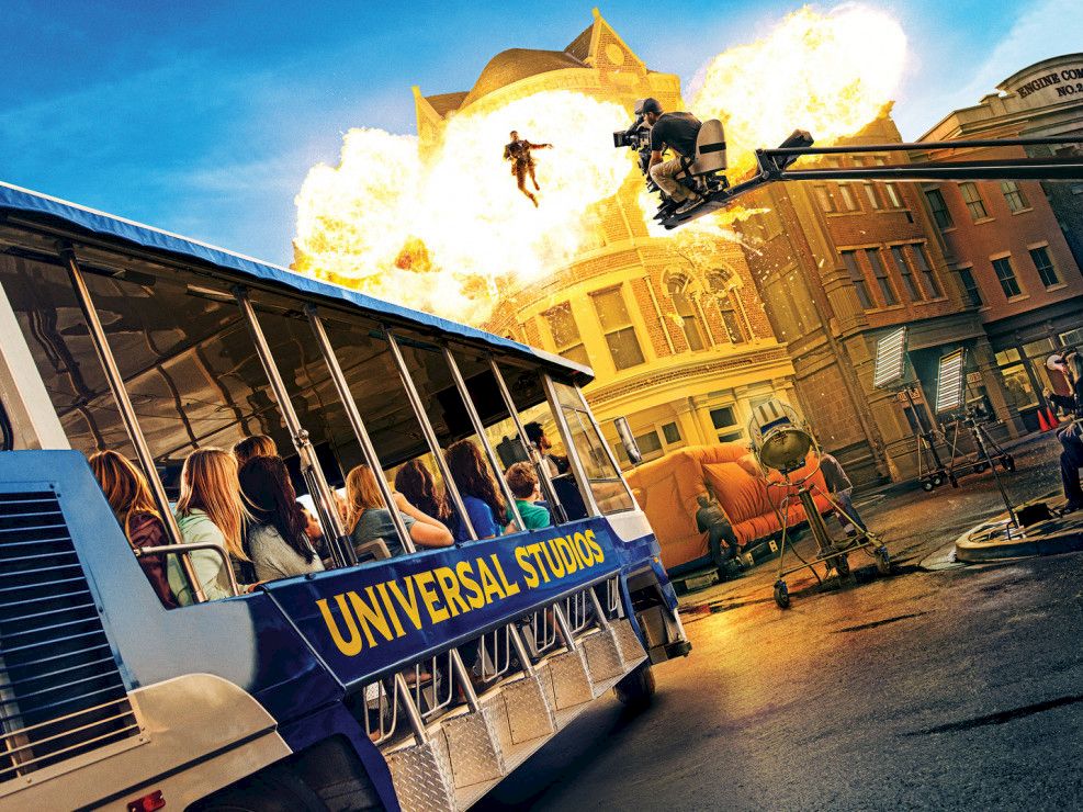 A Universal Studios tram drives through a stunt set as explosions and fire blast toward a brick building, with performers firing.