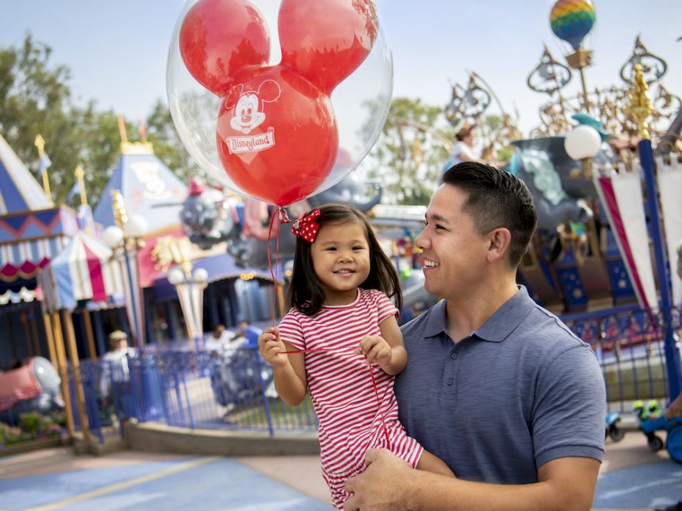 Disneyland day: a father holds his smiling toddler with a giant red Mickey balloon in a festive parade street.