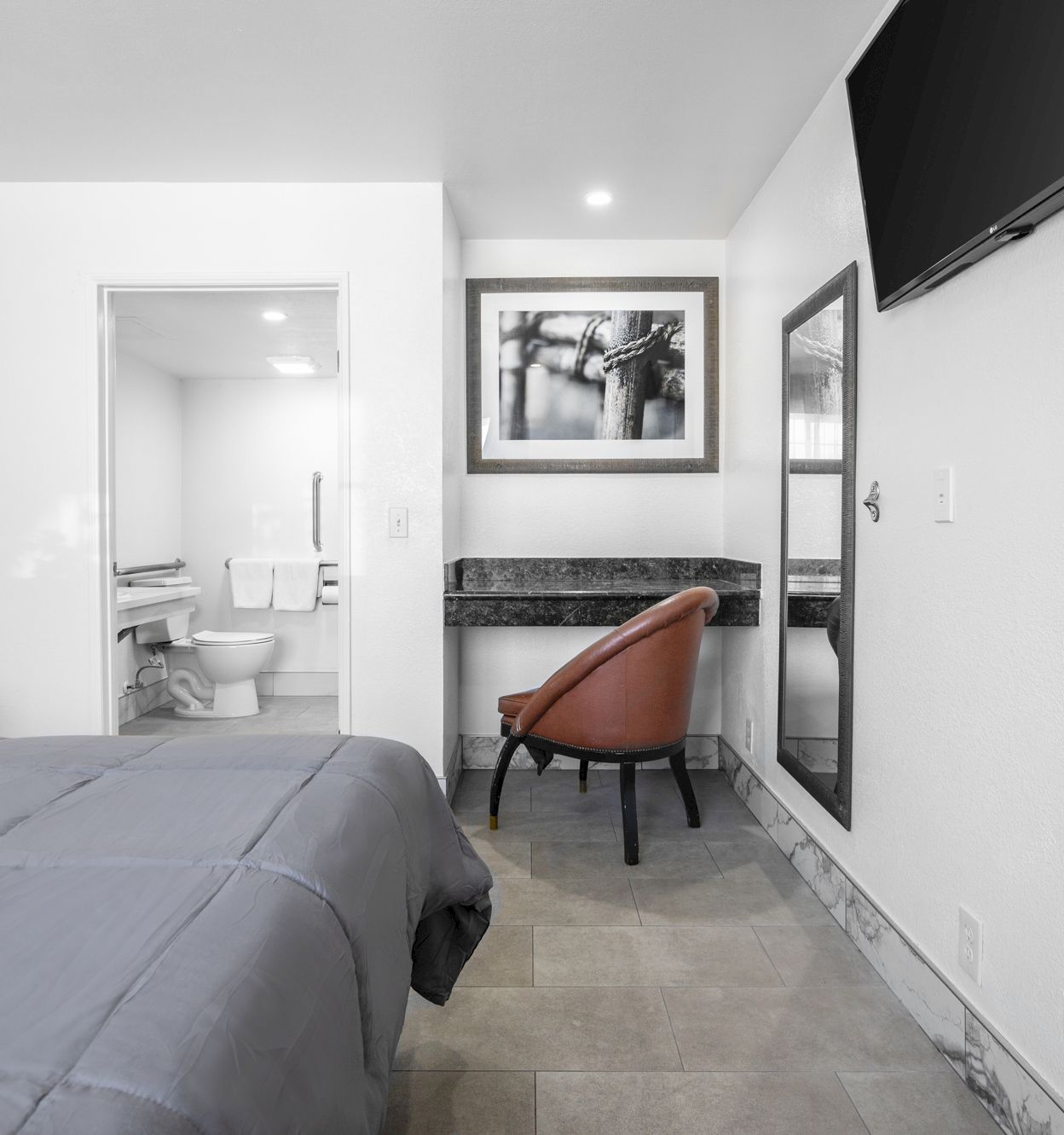 A modern bedroom with a bed, brown chair, wall TV, and framed photos, opening into a clean bathroom with toilet.