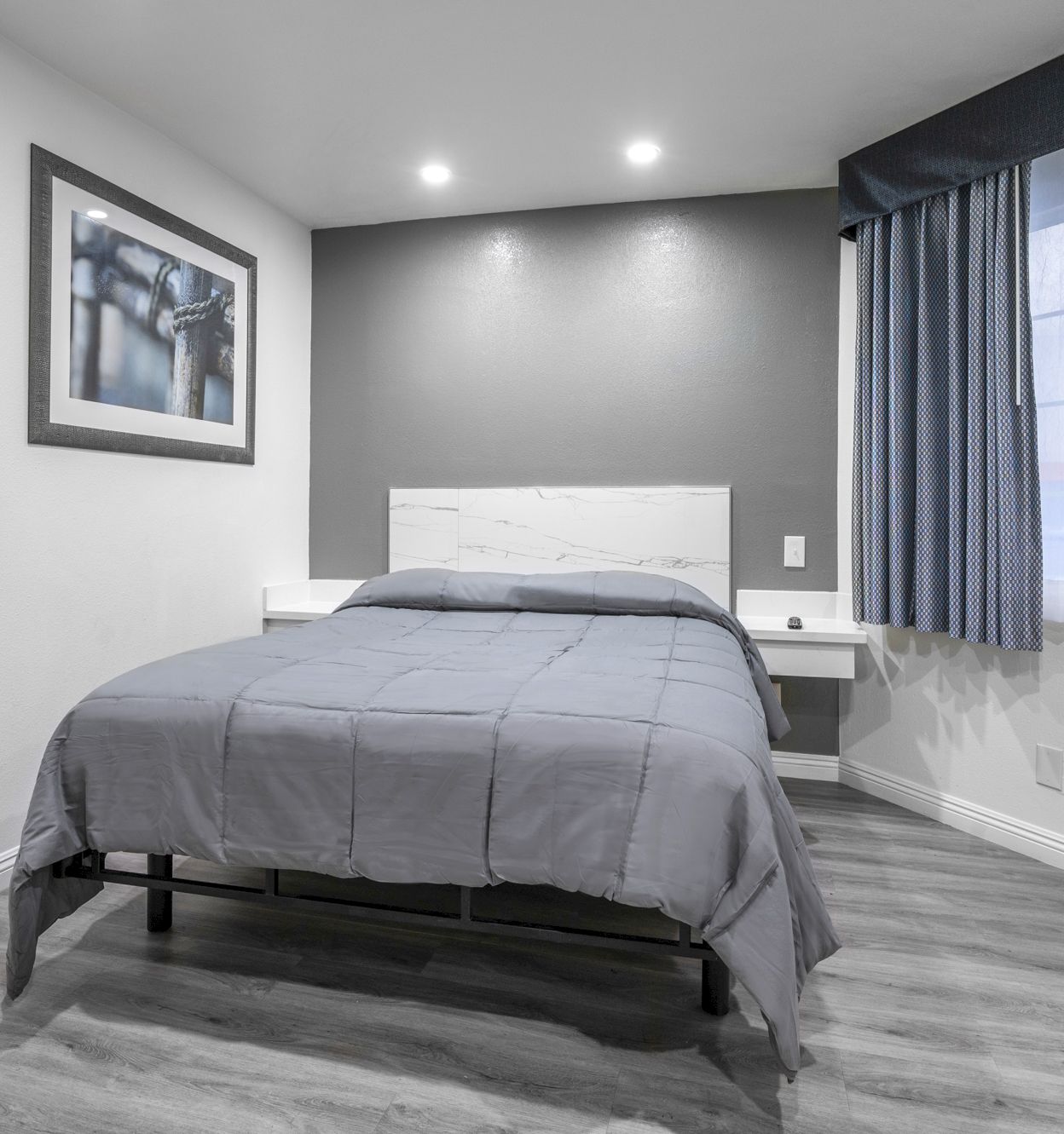 A modern bedroom with a gray bed, framed wall art, and blue-pattern curtains by a bright window.