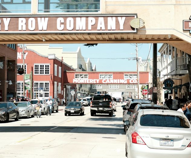 A sunny downtown street scene with parked cars and storefront signs reading &ldquo;ROW COMPANY&rdquo; and &ldquo;MONTEREY CANNING CO,&rdquo; among others.