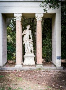A classical statue of a woman holding a child stands between two pink columns in a garden courtyard.