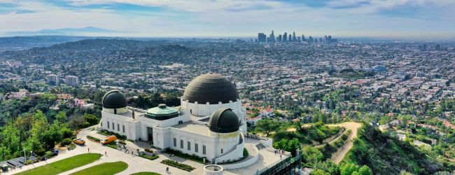 Aerial view of a white observatory with domes overlooking a sprawling city skyline beneath dramatic blue clouds.