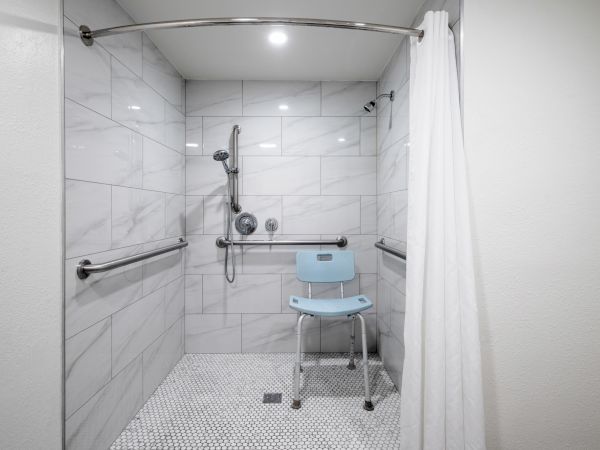 Accessible walk-in shower with marble tile, handheld showerhead, grab bars, a shower chair, and a white curtain.