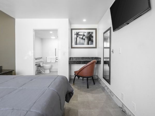 A modern bedroom with a bed, brown chair, wall TV, framed art, and a view into a white bathroom with toilet.