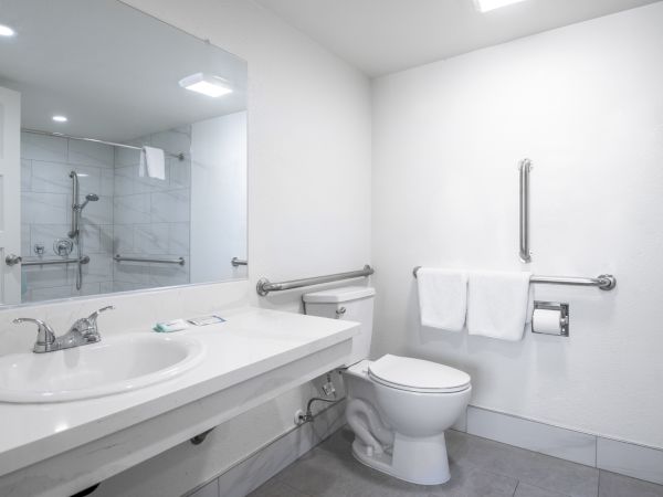Bright hotel bathroom with double sink, toilet, bathtub/shower, towels, and grab bars.