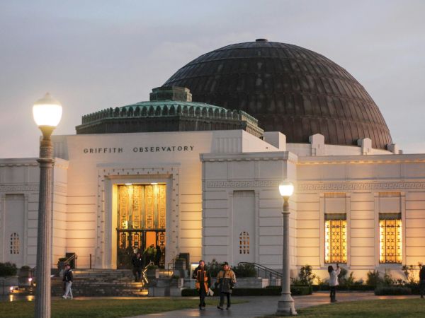 The Griffith Observatory dome rises above its building as people walk by at dusk.