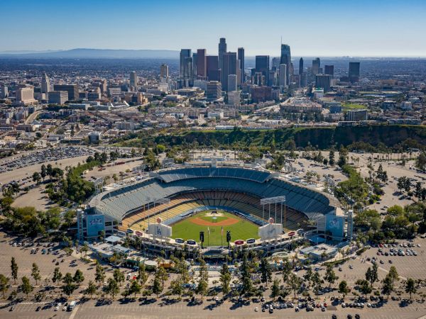 Aerial view of Dodger Stadium in Los Angeles with the LA downtown skyline towering in the background, under a clear blue sky.