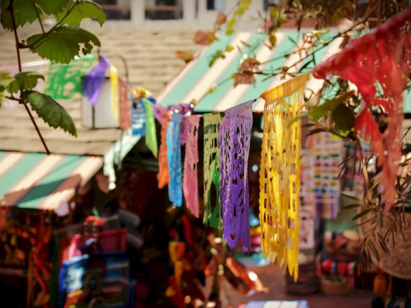 Colorful hanging festival decorations and banners sway in a courtyard, with green leaves in the foreground.