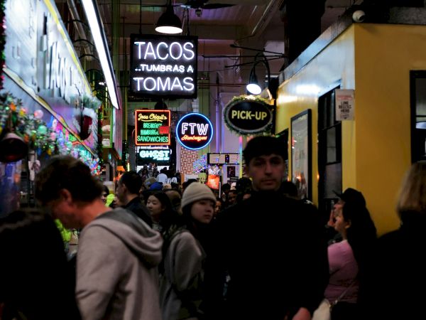 A crowded night street with neon signs for tacos and other fast-food spots, including &ldquo;TACOS&hellip; TOMAS,&rdquo; glowing overhead.