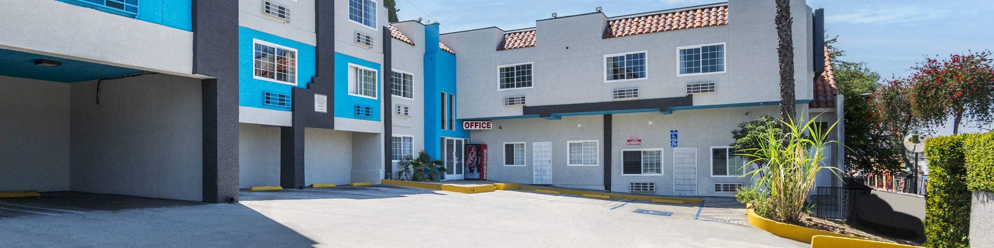 A sunny courtyard beside modern blue-and-white buildings, with windows, tiled roofs, and an &ldquo;OFFICE&rdquo; sign on the wall.