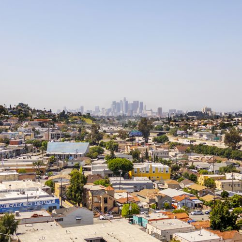 Aerial view over neighborhoods toward a distant downtown skyline under a clear blue sky.