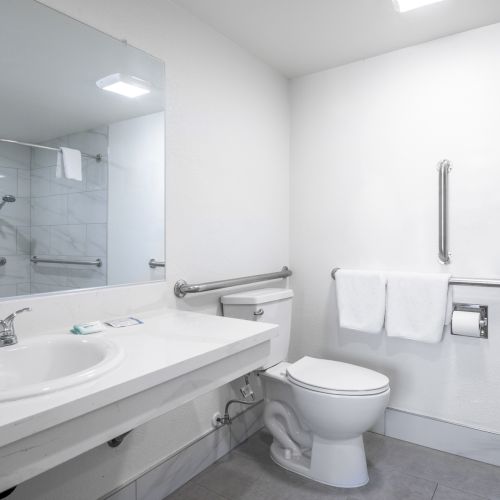 A bright modern bathroom with a vanity sink, toilet, and tub-shower combo with towels and grab bars.