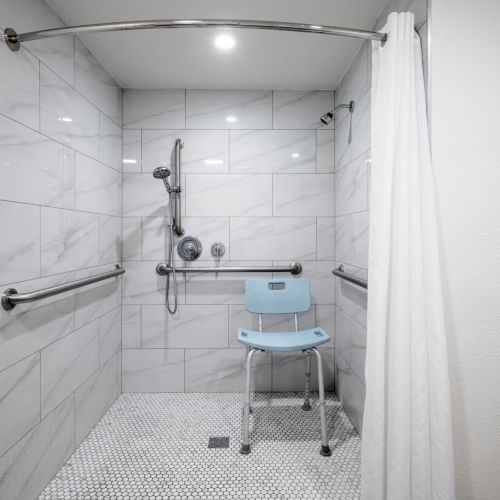 Accessible shower with marble tiles, handheld showerhead, grab bars, and a blue seat under a white curtain.
