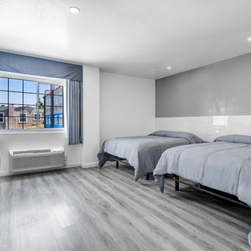 A bright bedroom with two beds, gray curtains, a window, an air conditioner, and a cushioned chair.