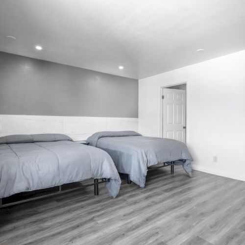 A bright modern bedroom with two beds, a grey accent wall, white door, and recessed ceiling lights.