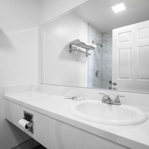 A bright bathroom with a white sink, countertop mirror, and shower/tub area behind a glass wall.